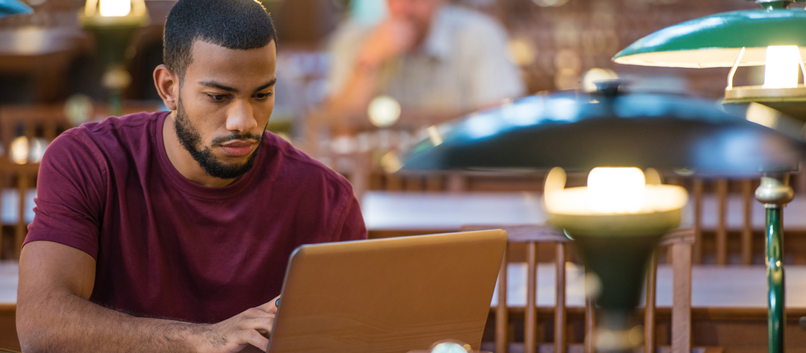 An African-American students sits at a table in a library, working on a laptop.